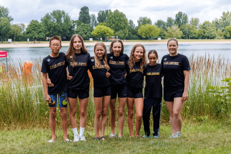 Gruppe junger Sportler in schwarzen T-Shirts mit „Karlsruhe“ am Wasser, umgeben von grüner Landschaft.