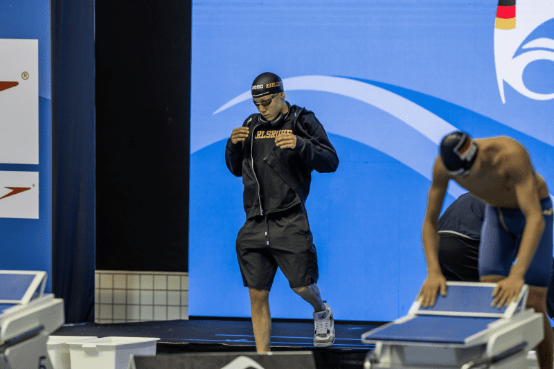 Ein Schwimmer in schwarzer Trainingskleidung betritt die Wettkampfbühne mit einem Handtuch in der Hand.