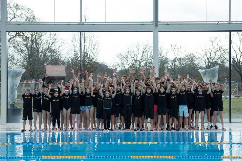 Gruppe von Schwimmern in schwarzen T-Shirts, die am Beckenrand jubeln, mit Glaswänden und Außenblick im Hintergrund.